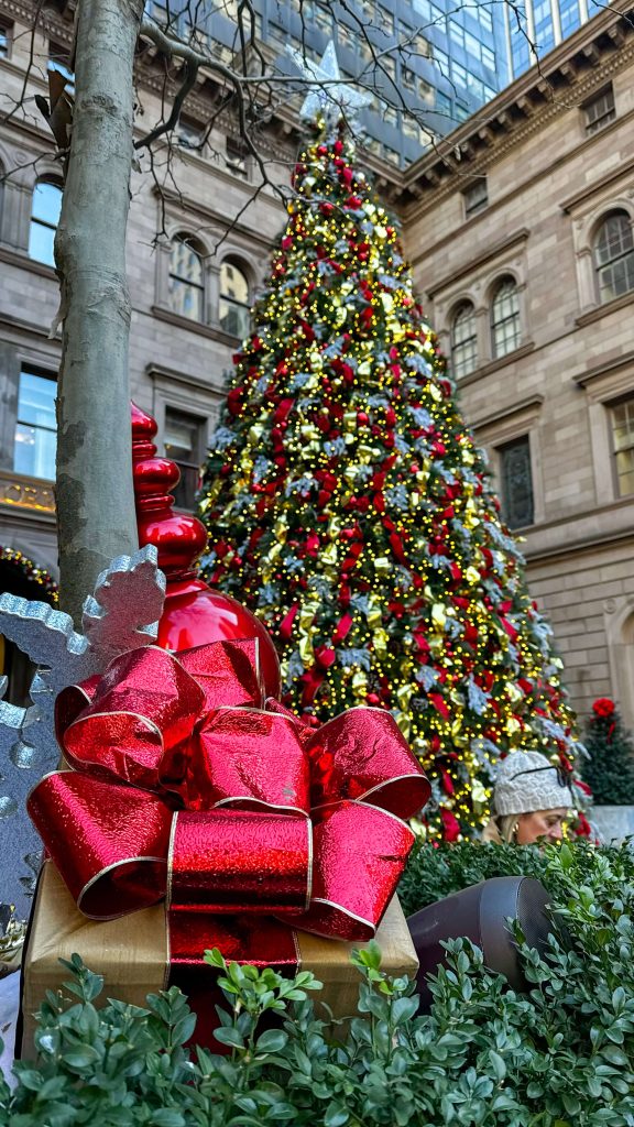 A towering Christmas tree at the Lotte New York Palace in Midtown Manhattan sparkles with warm white lights, red ribbon garlands, gold ornaments, and a glowing star topper. A large wrapped gift with a shiny red bow sits in the foreground, highlighting the festive holiday displays that make this stop a favorite on an NYC Christmas Trip.