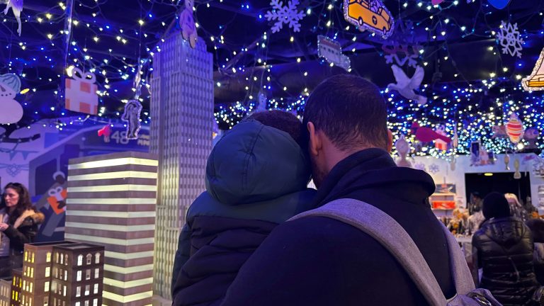 parent carries a child through a festive indoor display filled with blue and white Christmas lights and miniature New York City buildings. This magical holiday scene is a memorable stop for an NYC Christmas itinerary with kids.